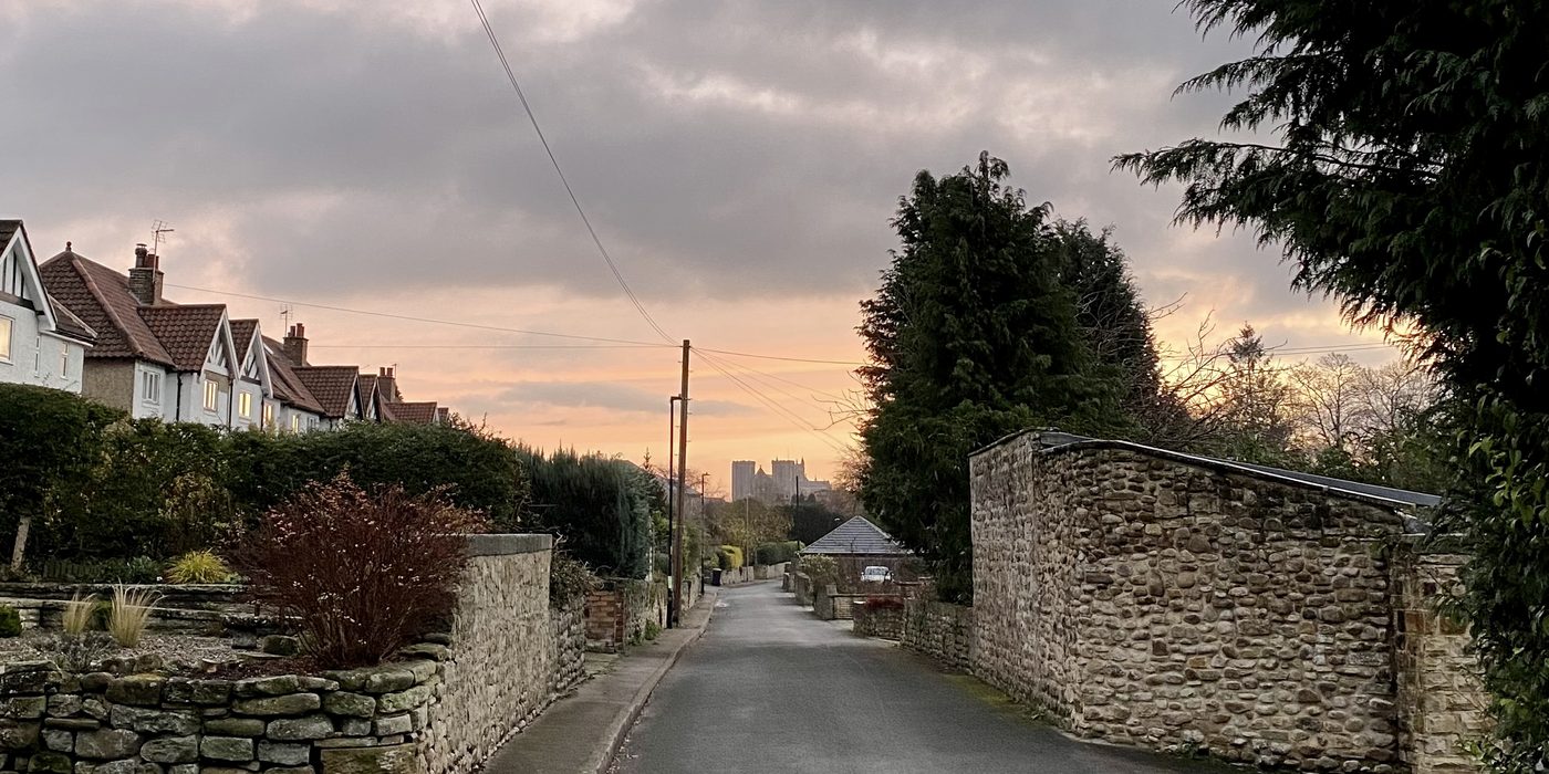 Ripon Cathedral seen at dusk from a residential street, framed by drystone walls and rendered semi-detached houses.